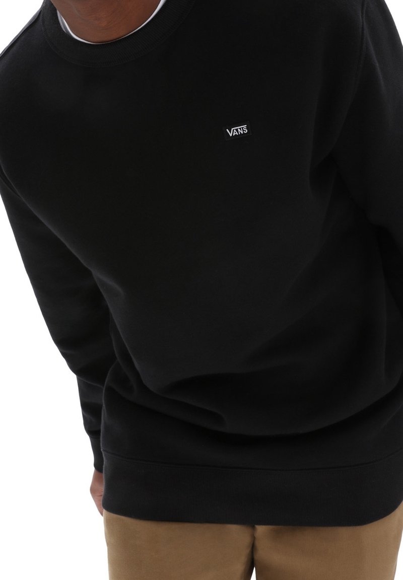 Personne portant un sweat-shirt Vans noir avec un petit logo blanc sur la poitrine et un pantalon marron clair, penchée légèrement en avant.