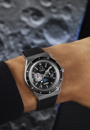 Montre-bracelet Timex avec bracelet en cuir noir, cadran noir, logos NASA et phase lunaire, date et sous-cadrans affichant le jour et les secondes.