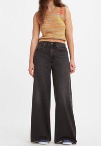 Woman wearing multicolored sleeveless knit top, high-waisted wide-leg black jeans, and blue sneakers standing against white background.