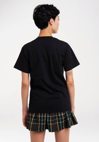 Schwarzes T-Shirt mit kurzen Ärmeln, kombiniert mit einem gefalteten, grün karierten Rock mit roten und gelben Akzenten, von hinten gesehen.