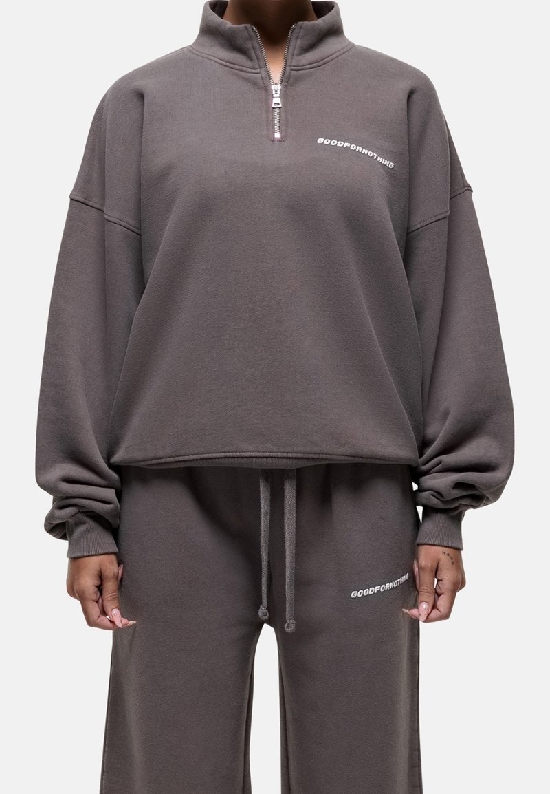 Personne portant un survêtement gris ample avec un sweat à demi-zip et un pantalon assorti, les deux marqués "GOODFORNOTHING" en lettres blanches.