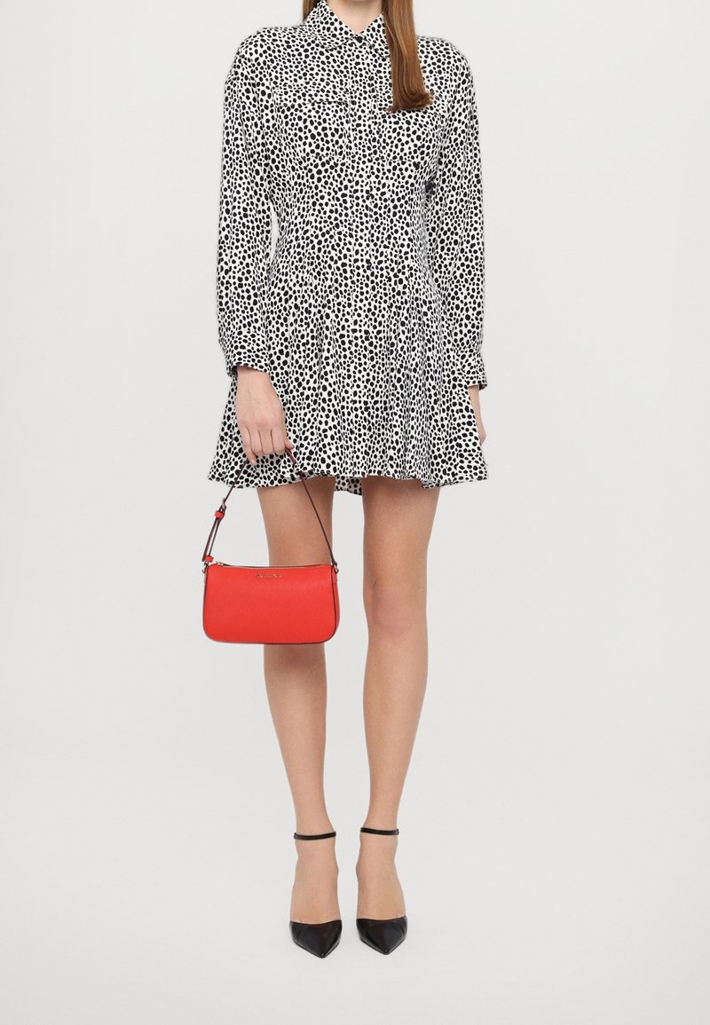 Femme portant une robe blanche avec des taches noires, des talons noirs pointus, et tenant un petit sac à main rouge par sa bandoulière.