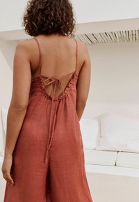 Jumpsuit od lanenog materijala boje hrđe s niskim leđima, nabranom teksturom i prilagodljivim vezicama. Glatka površina i ležeran kroj.