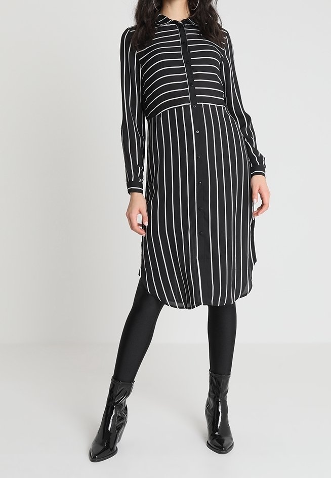 Femme portant une robe midi boutonnée à rayures verticales noires et blanches, des leggings noirs et des bottines noires brillantes, debout devant un fond uni.