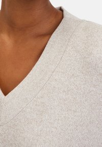 Gros plan sur un cou et un haut de poitrine à la peau foncée portant un pull en tricot beige à col V avec une texture subtile.