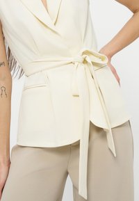 Γυναίκα που φοράει μπεζ αμάνικο μπλέιζερ δεμένο με φιόγκο στη μέση και μπεζ παντελόνι, με ορατά τατουάζ στο μπράτσο.