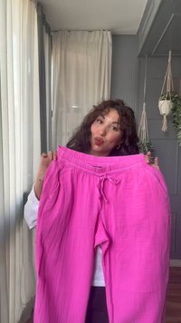 Femme tenant un pantalon large rose vif avec cordon dans une pièce avec des plantes suspendues et des rideaux transparents.
