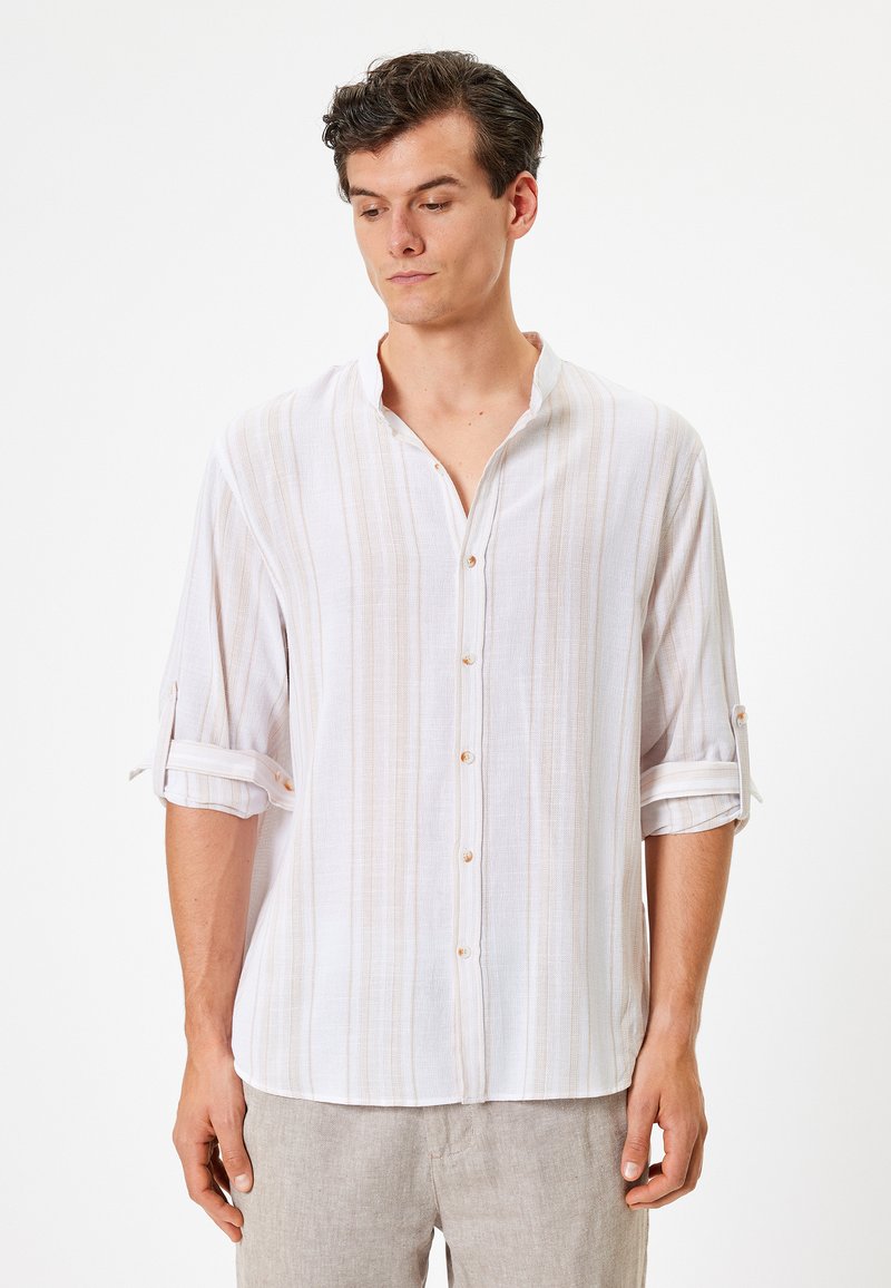 Valkoinen nappikaulusshirt, jossa on vaaleat pystysuorat raidat; valmistettu kevyestä kankaasta; käärittävät hihat, jotka on kiinnitetty nappien avulla.