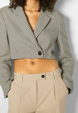 Vrouw die een kort geruit colbert draagt met één knoop en beige high-waist broek met handen in de zakken.