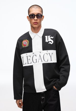 Mann trägt ein schwarz-weißes Langarmshirt mit dem Schriftzug "LOST LEGACY", einem Wappenemblem und Sonnenbrille, posiert mit einer Hand in der Tasche vor weißem Hintergrund.