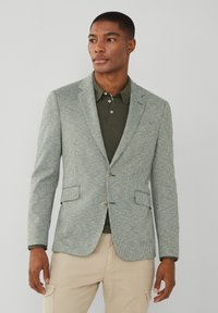 Svetlozelený blazer s houndstooth vzorom, s klopovými goliermi, dvoma prednými vreckami, zapínaním na jeden gombík a textúrovanou tkaninou, vrstvený na tmavozelenom polo.