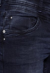 Gros plan sur la poche d'un jean en denim bleu foncé avec des coutures visibles, des rivets en métal et un détail de passants de ceinture.