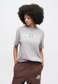Γκρι βαμβακερό T-shirt με στρογγυλή λαιμόκοψη, με μεγάλο λογότυπο "NIKE SHOX" σε λευκό και μπλε. Συνδυασμένο με καφέ φόρμες.