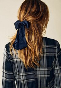 Laivastonsininen silkkinauha solmittuna rusetille, kiinnittäen aaltoilevat, vaaleanruskeat hiukset. Kuvioitu tumma ruudullinen paita, jossa näkyy harmaita ja laivastonsinisiä sävyjä.
