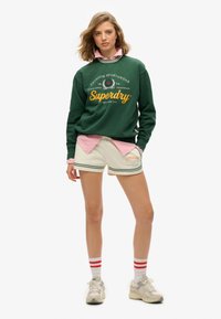 Sudadera verde con logo blanco, camisa de color rosa con cuello debajo, pantalones cortos beige con rayas verdes y zapatillas blancas con acentos rojos.