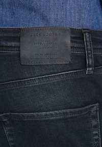 Jeans de mezclilla azul oscuro con un parche de cuero de la marca que dice "Jack & Jones. Jeans Intelligence." Presentan un diseño clásico de cinco bolsillos y costuras.