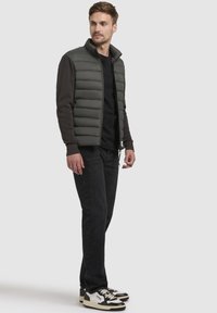 Grå polstret vest med mørke ærmer, lynlåslukning foran, båret over en sort skjorte og parret med sorte jeans og sneakers.