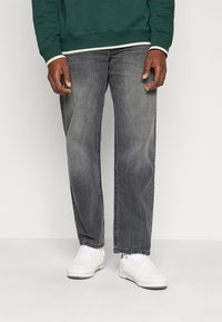 Jeans in denim grigio con taglio dritto, indossati con una felpa verde con bordi bianchi e sneakers bianche con accenti testurizzati.
