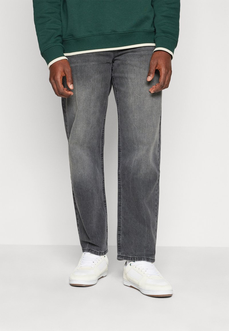 Jeans in denim grigio con taglio dritto, indossati con una felpa verde con bordi bianchi e sneakers bianche con accenti testurizzati.
