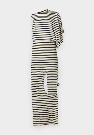 Femme portant une robe rayée noire et blanche à une épaule avec une fente haute sur la cuisse et des sandales jaunes, debout devant un fond gris uni.