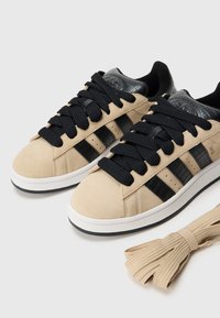 Μπεζ σουέτ αθλητικά παπούτσια με μαύρες δερμάτινες λεπτομέρειες, χοντρούς μαύρους κορδόνια και τρεις μαύρες ρίγες στα πλάγια. Περιλαμβάνει ένα επιπλέον ζευγάρι μπεζ κορδόνια.