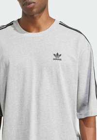 T-shirt grigio a maniche corte realizzato in tessuto morbido. Presenta un design a tre strisce nere sulle spalle e un piccolo logo Adidas nero sul petto.