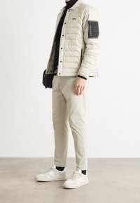 Beige vadderad jacka med svarta accentfärger, slim-fit ljusbeiga byxor och vita sneakers med grå och svarta detaljer. Mjuk textur, modern design.