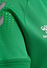 Grön tröja med ett strukturerat tyg, med cirkulära mönster. Grå detaljer och en vit logotyp på bröstet. Korta ärmar.