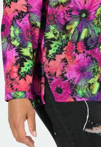 Farverig blomsterbluse med en blanding af pink, grønne og sorte nuancer, lavet af et let stof og med oprullede ærmer. Kombineret med sorte jeans.