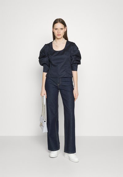 Γυναίκα που φοράει μπλούζα με ναυτικό φούσκα μανίκι, σκούρες φαρδιές τζιν, λευκά sneakers, ένα στρώμα από μαργαρίτες και κρατάει μια λευκή τσάντα με αλυσίδα.