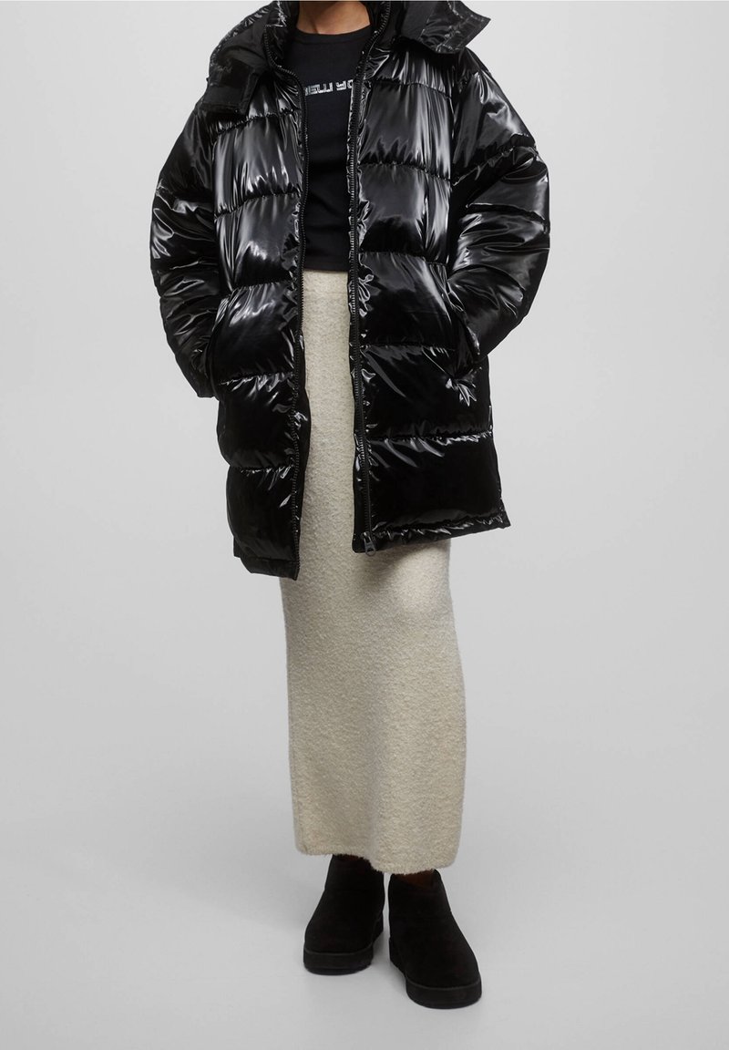 Manteau matelassé noir brillant avec capuche, motif matelassé, porté sur une chemise noire et une jupe en maille crème, assorti à des bottines noires.