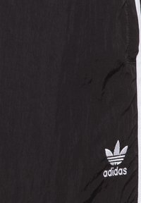 Pantalons légers en polyester noir avec une texture lisse, des rayures blanches sur les côtés et un logo Adidas en fil argenté brodé près de l'ourlet.