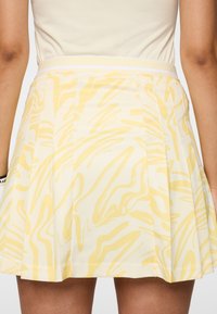 Femme portant une jupe plissée blanche avec un motif abstrait jaune clair et une ceinture blanche assortie.