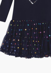 Abito blu navy con maniche lunghe, caratterizzato da un collo decorato con applicazioni di paillettes. Gonna in tulle, abbellita da punti riflettenti multicolori e orlo arricciato.
