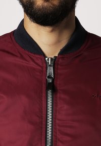 Burgunderröd bomberjacka med dragkedja fram, ribbat marinblått krage och broderad logodetalj. Slät textur, lätt material.