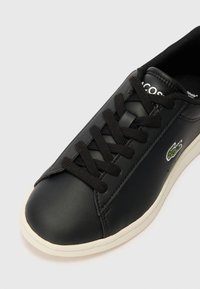 Sneaker in sintetico nero con lacci neri, punta rotonda e suola beige. Presenta un piccolo logo verde ricamato sul lato.
