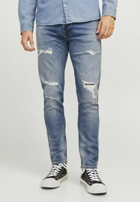 Világoskék farmernadrág, melynek elülső részén kipolírozott szakítások találhatók, és slim fit szabású. Fekete sportcipővel és fehér fűzőkkel párosítva.