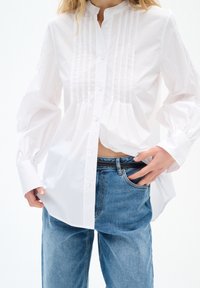 Personne portant une chemise blanche à manches longues boutonnée avec des plis et un jean bleu, une main dans la poche et l'autre tenant l'ourlet de la chemise.
