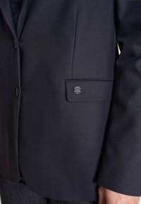 Merihenkinen bleiseri, joka on valmistettu sileästä kankaasta ja jossa on yksittäinen napin suljenta, sivutasku, jossa on pyöreä musta logo, sekä sopiva leikkaus.