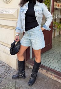 Lichtblauwe denimjas en korte broek, zwarte geribbelde coltrui, zwarte kniehoge laarzen, een zwarte gevlochten handtas vasthoudend. Getextureerde materialen.