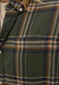 Close-up of olive green plaid shirt with navy, brown, and white stripes, showing a chest pocket and button details.