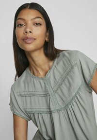 Jasnozielona bluzka wykonana z miękkiego materiału, z koronkowym wykończeniem, krótkimi rękawami i plisowanymi detalami z przodu. Okrągły dekolt.