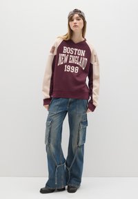 Bordeaux sweatshirt met de tekst "BOSTON NEW ENGLAND 1998", beige mouwen; blauwe cargobroek met meerdere zakken; zwarte schoenen; grijze pet.