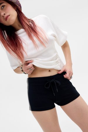 Ung person med langt lyserødt hår iført en hvid ruched crop top, sorte shorts med snøre og en ternet hat, poserer mod en ensfarvet baggrund.