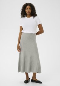 Femme aux cheveux bouclés et foncés portant une chemise blanche à manches courtes, une jupe midi gris clair et des ballerines noires, se tenant devant un fond uni.