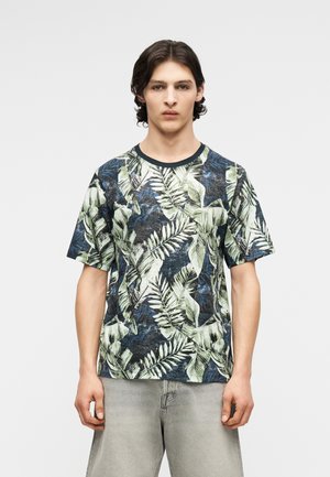 Mladý muž v krátkym rukáve so vzorom tropických listov v odtieňoch zelenej, modrej a čiernej, v kombinácii so svetlosivými nohavicami.