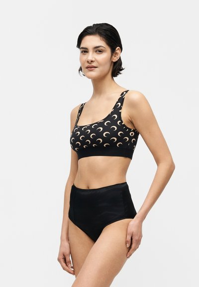 Γυναίκα με κοντά σκούρα μαλλιά, που φοράει μαύρο crop top με σχέδια ημισελήνου και μαύρο ψηλόμεσο εσώρουχο, στέκεται μπροστά σε λευκό φόντο.