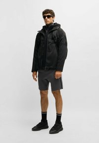 Svart vattentät jacka med huva, dragkedjefickor, bärs över en svart polotröja och grå shorts. Svarta sneakers och strumpor med logotypdetaljer.