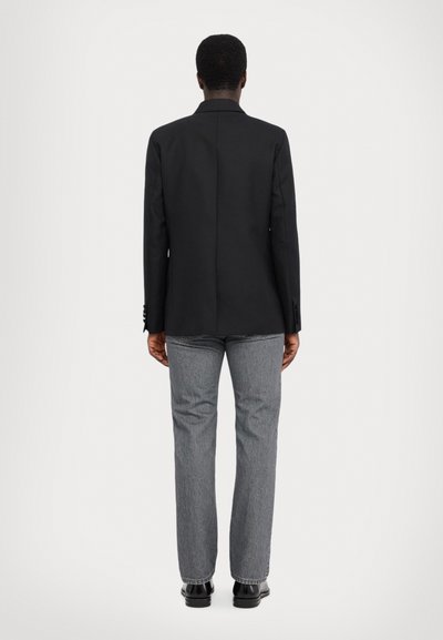 Zwarte blazer met een getailleerde pasvorm, enkele split en knoopmanchetten, gecombineerd met grijze straight-leg jeans en zwarte schoenen.
