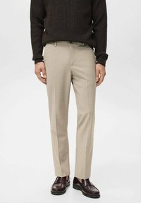 Pantalones beige de corte recto con una textura suave, presentan un frente plano, bolsillos laterales y una pierna ajustada, combinados con un suéter oscuro texturizado y zapatos.